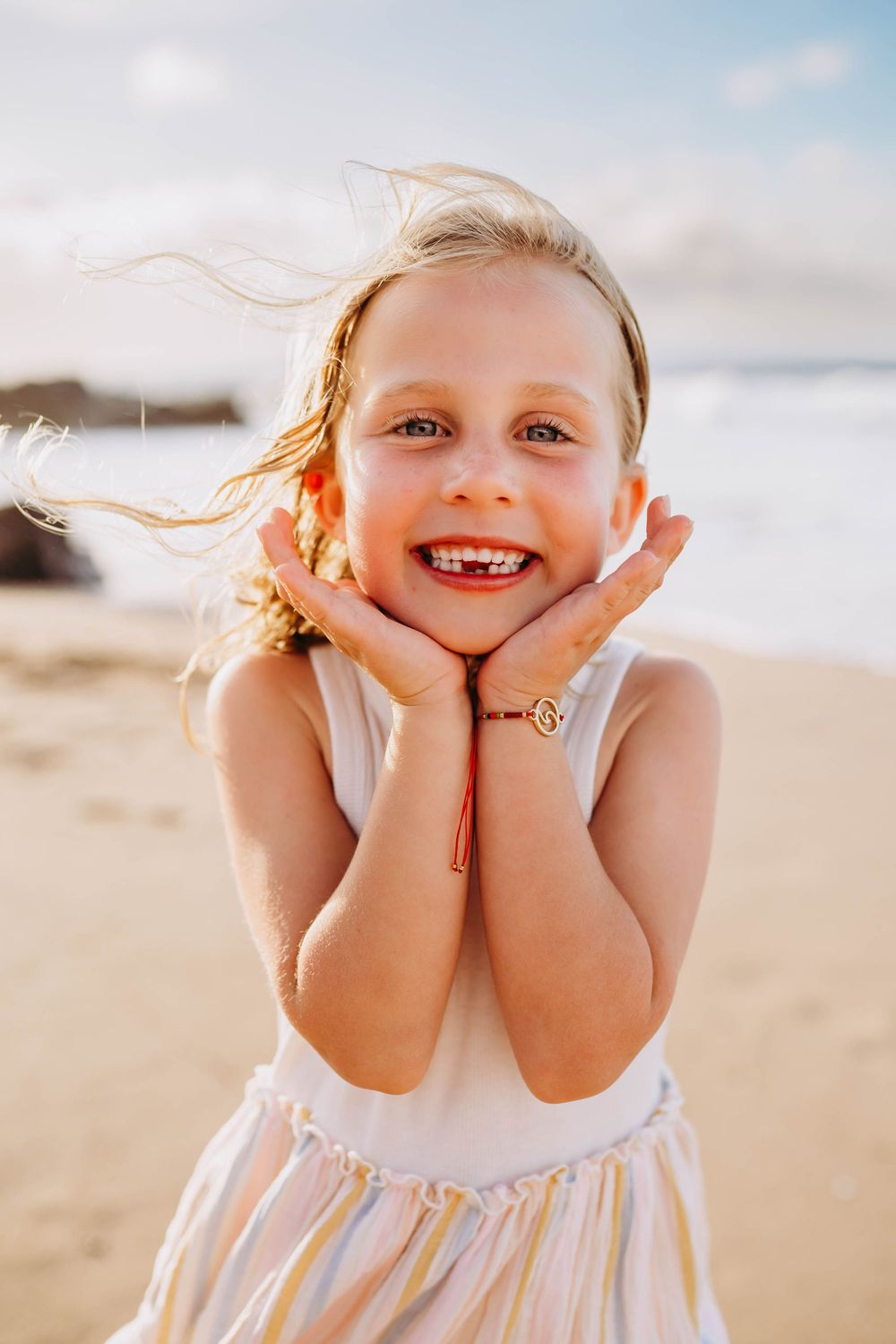 A joyful child poses on a sunny beach wearing a white dress as their hair blows in the ocean breeze.
