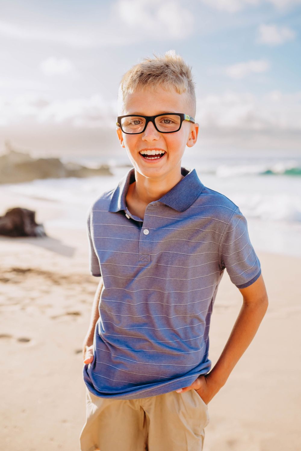 Young boy in glasses and blue polo shirt laughing and playing on sunny beach.