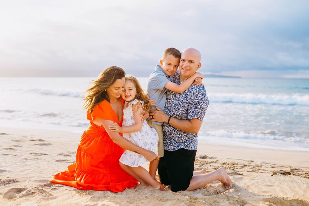A family shares a joyful moment together while kneeling on a sunny beach with ocean waves in the background.