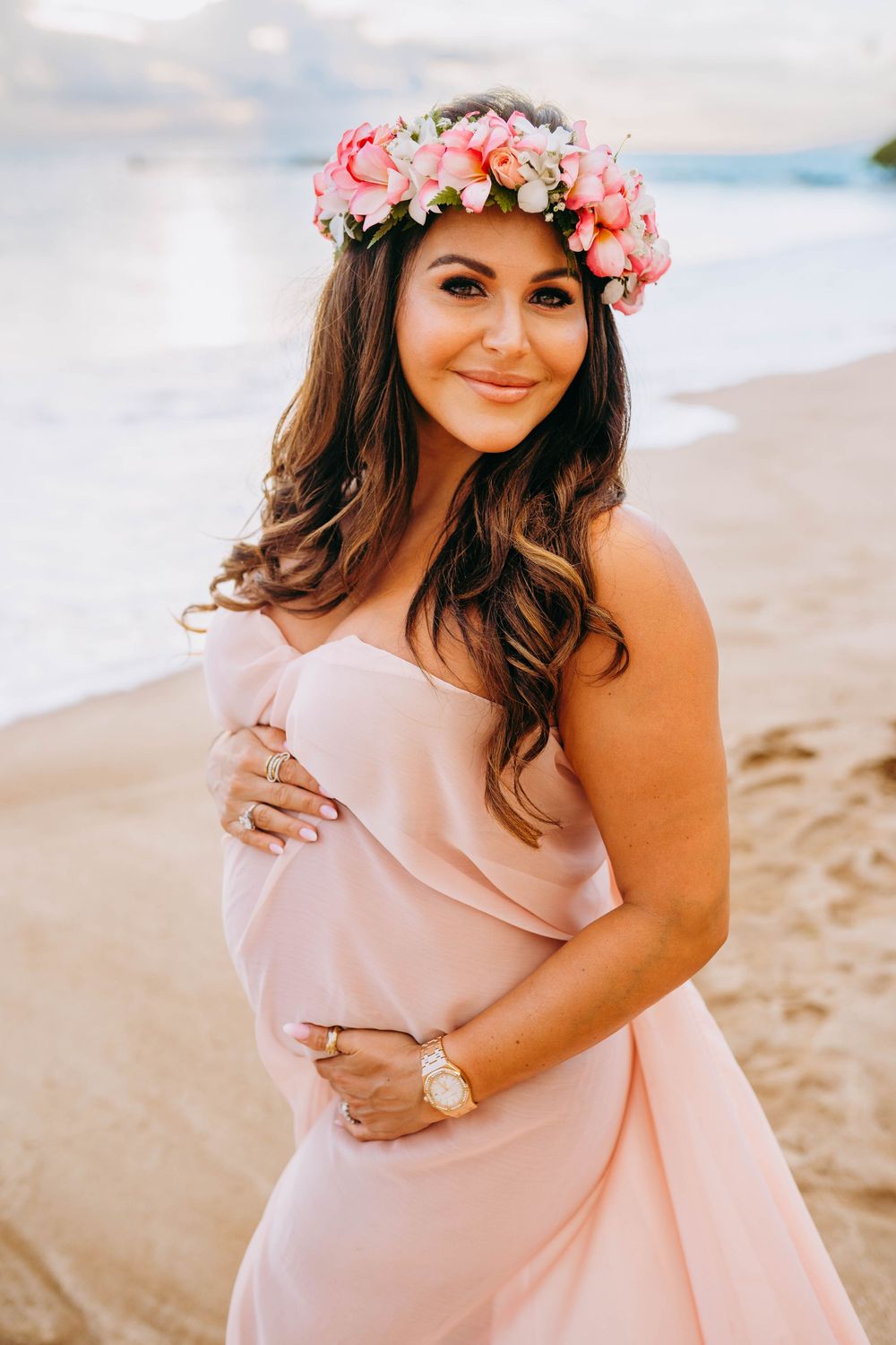 A person wearing a pink floral crown and blush-colored dress poses on a beach at sunset.