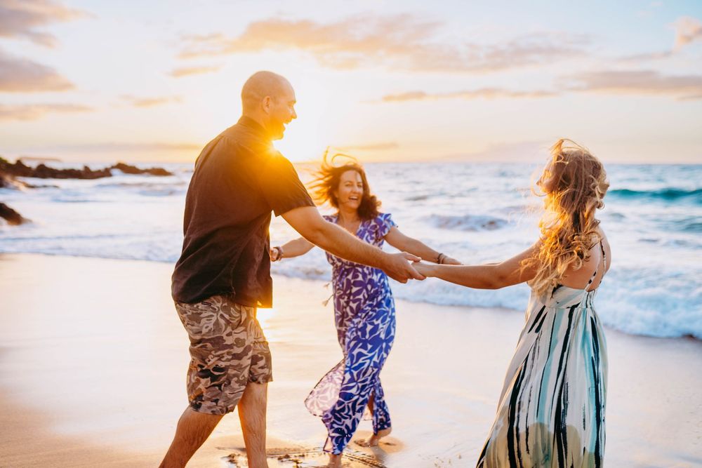 Three people hold hands and dance together on a beach at sunset as ocean waves crash behind them.