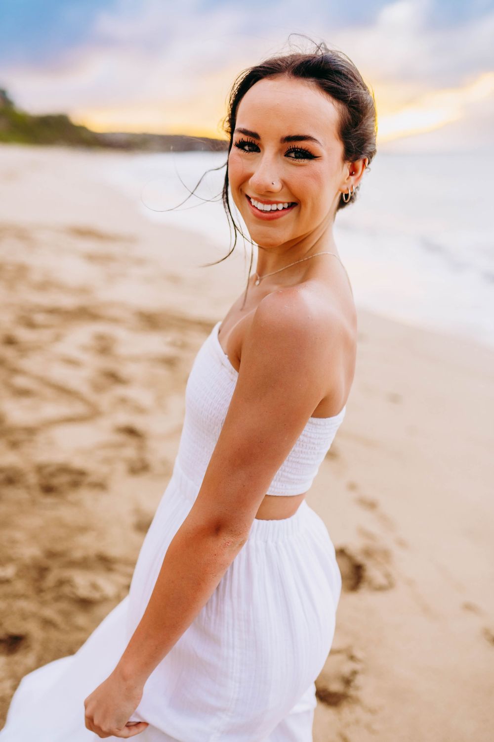 A person in a white sundress poses joyfully on a sandy beach at sunset with soft lighting and ocean views.
