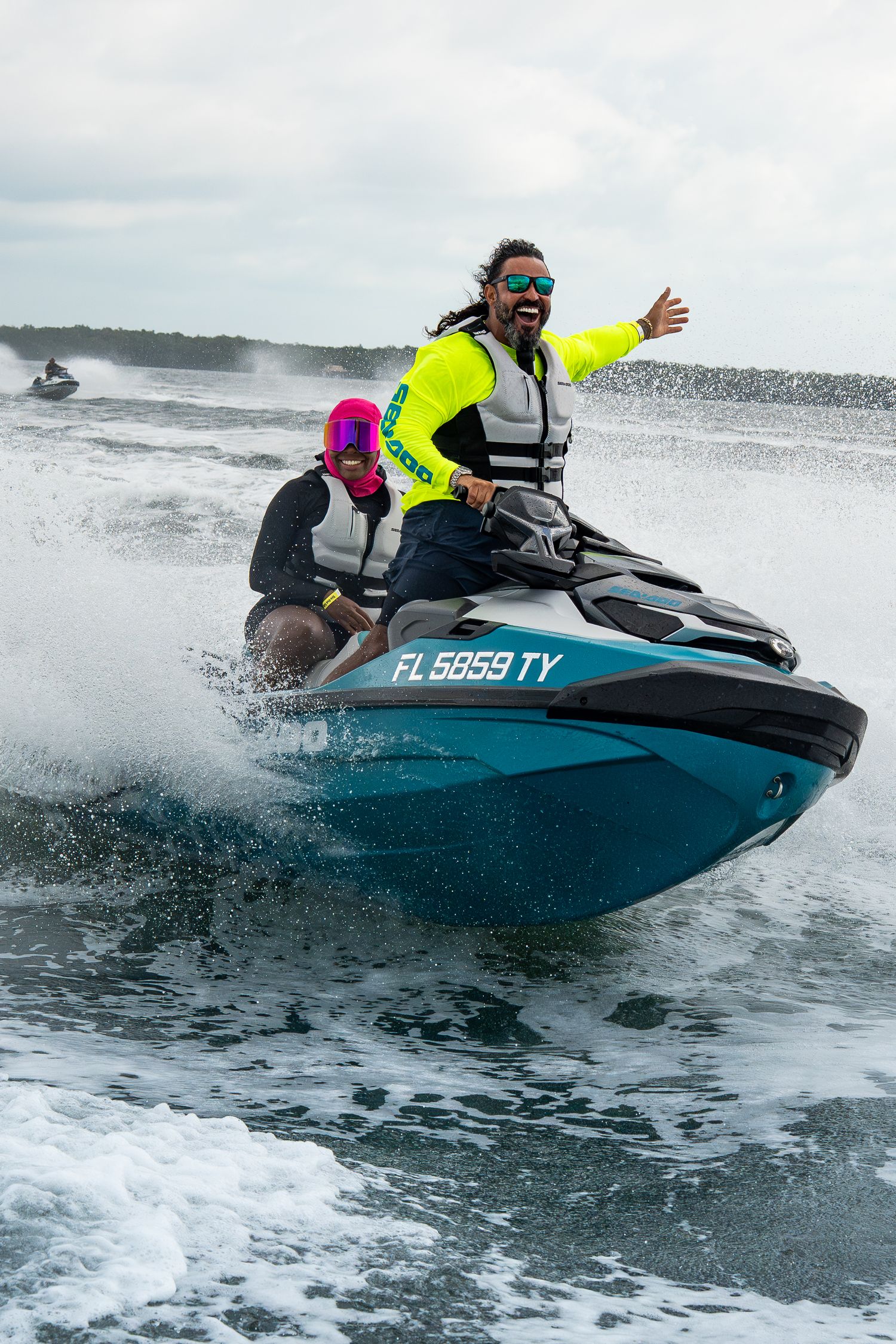 Two people riding a blue jet ski over ocean waves while wearing safety gear and bright neon clothing.