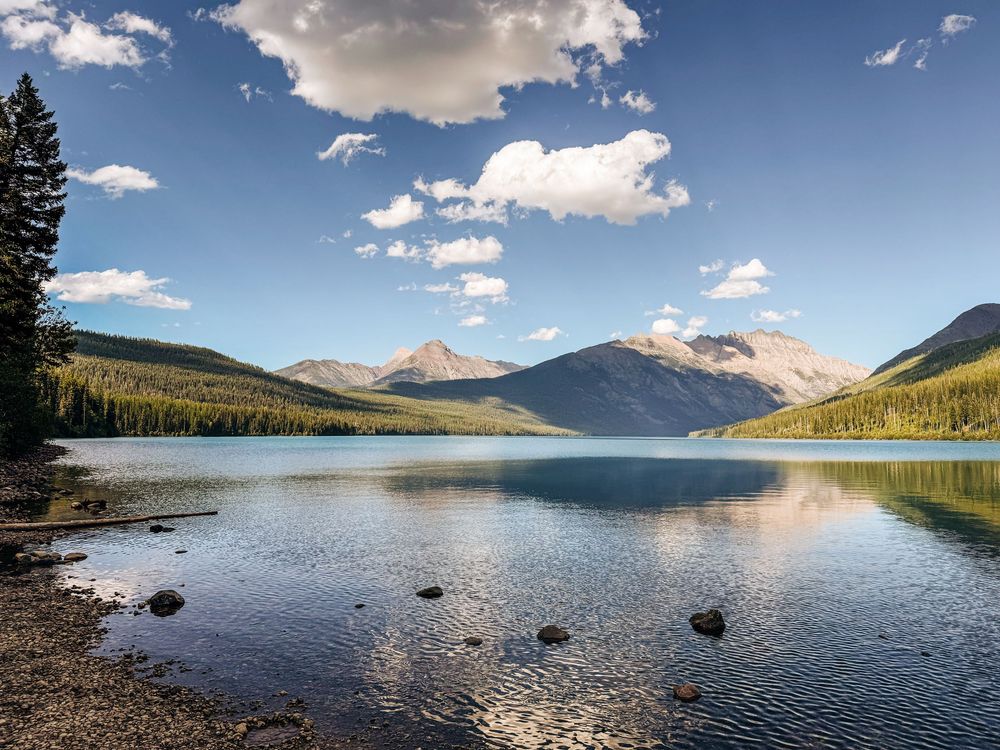 Scenic mountain lake with calm waters reflecting blue sky and puffy clouds surrounded by evergreen forest slopes.