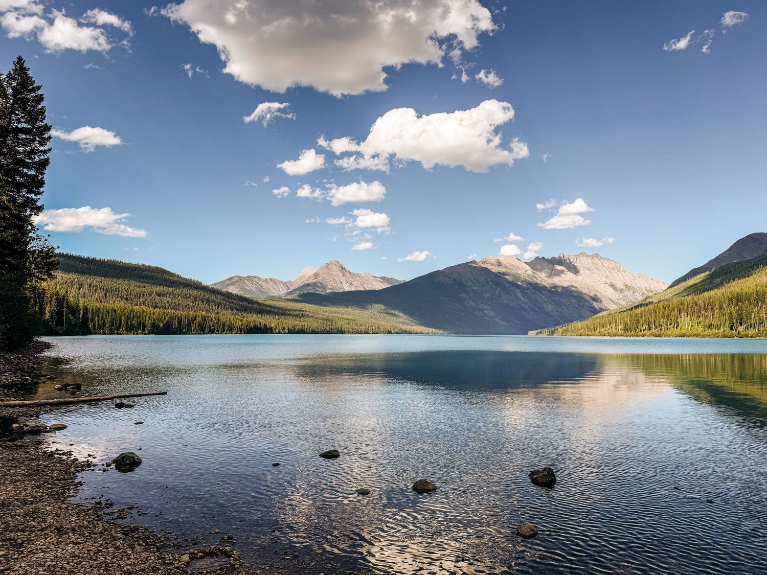Scenic mountain lake with calm waters reflecting blue sky and puffy clouds surrounded by evergreen forest slopes.