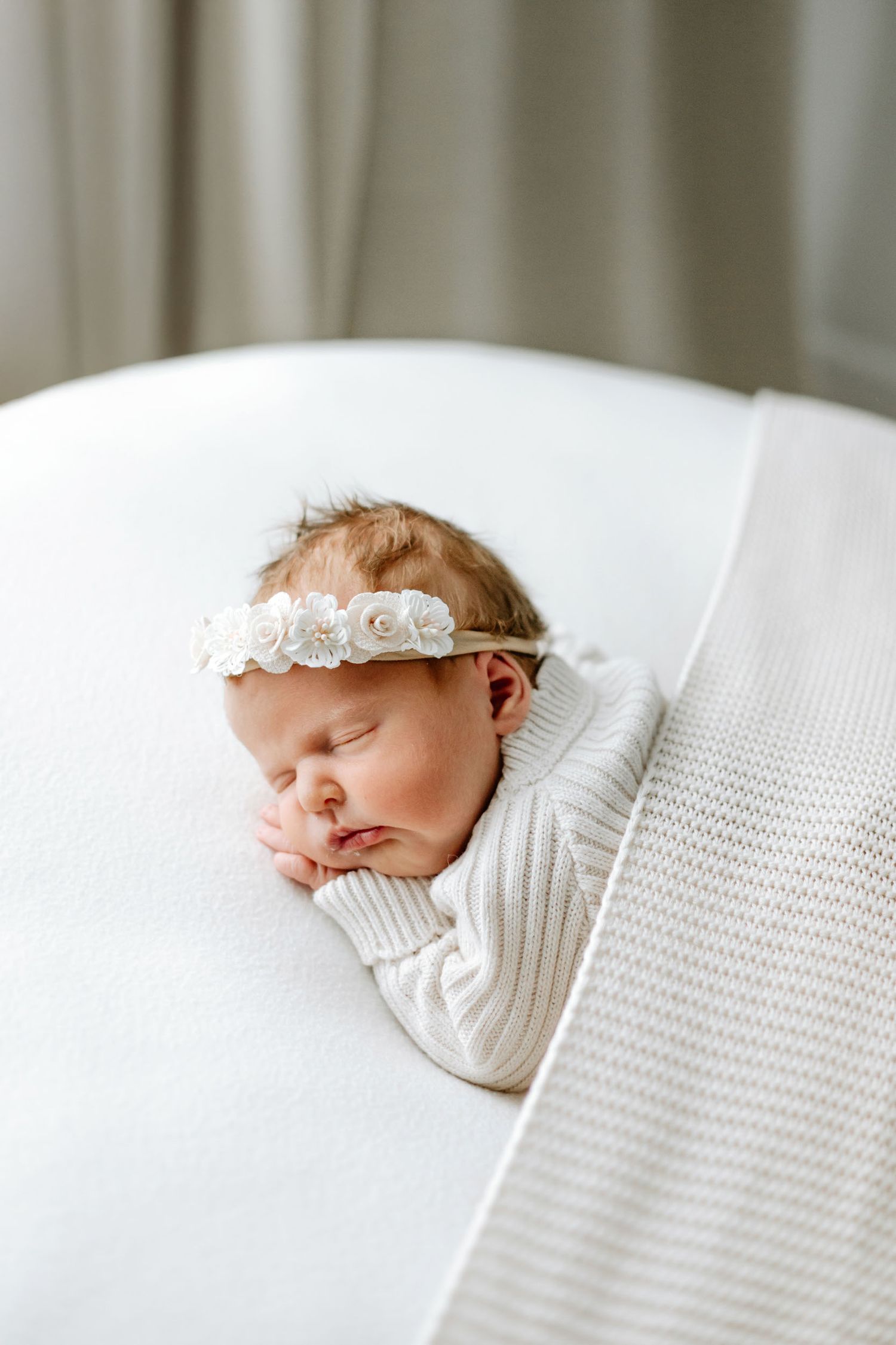 Sleeping newborn baby wearing white floral headband rests peacefully on textured white bedding.