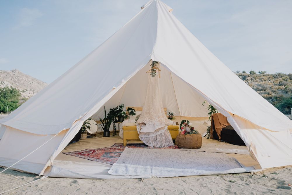 White bell tent glamping setup in desert landscape with boho rugs, yellow cushions, and potted plants.