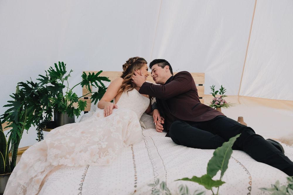 Bride and groom share a romantic moment lying together on a bed inside a boho bell tent surrounded by lush greenery.
