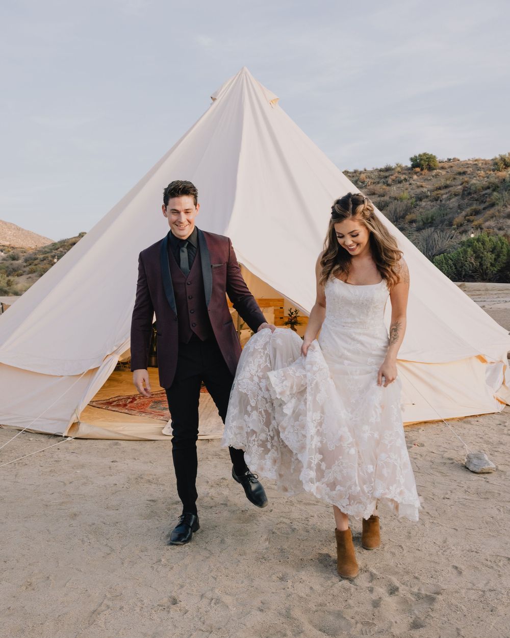 Bride and groom walk hand in hand in front of a white bell tent in a desert landscape.