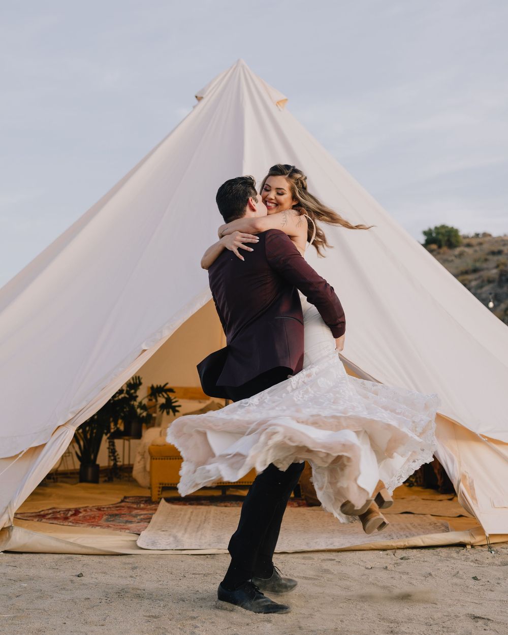 A groom lifts his bride in a white lace wedding dress outside a large white bell tent at a boho outdoor wedding.