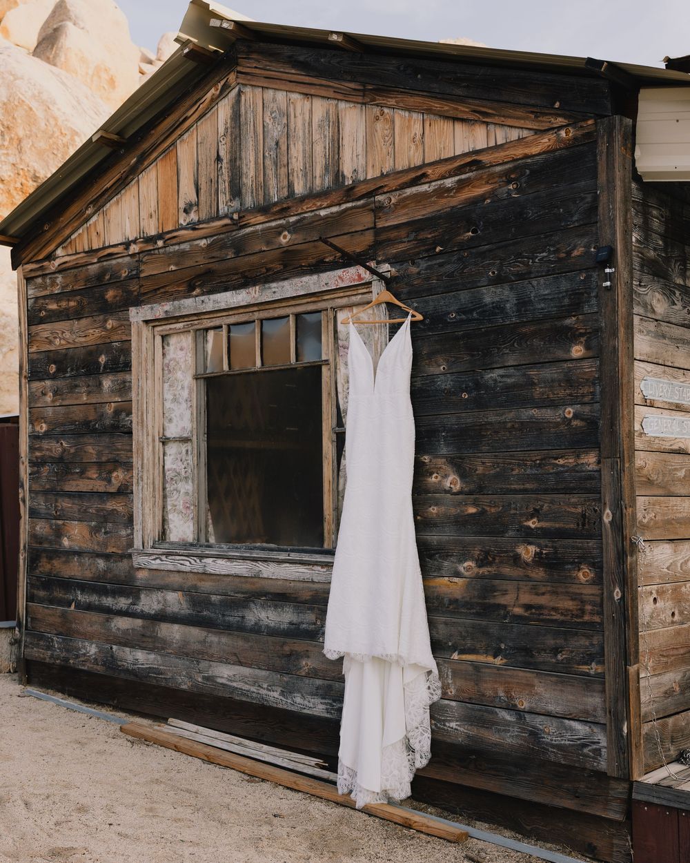Elegant white wedding dress hanging on rustic weathered wooden cabin exterior in desert setting.