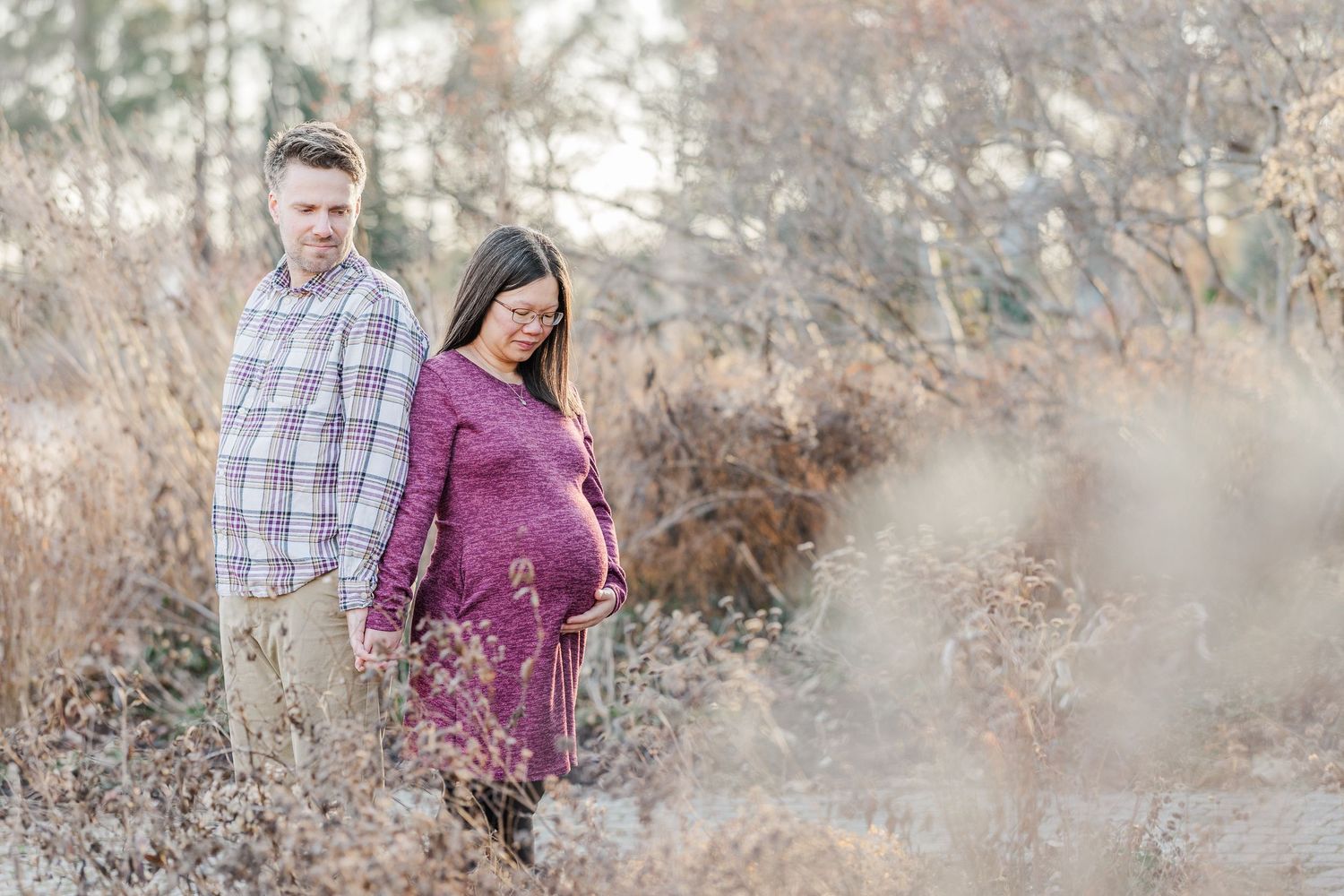 A couple stands together in a field of dried winter grass, with the expectant mother wearing a purple maternity dress.