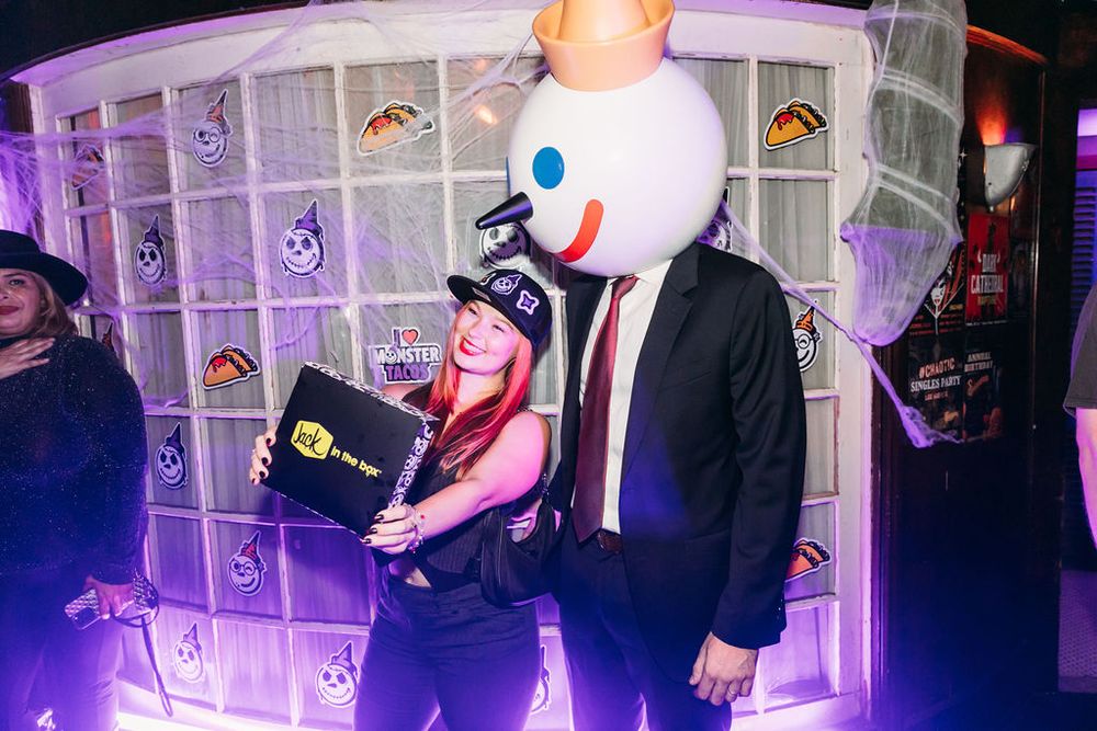 A person in a black suit and large white mascot head poses with someone in pink hair at a nightclub event.