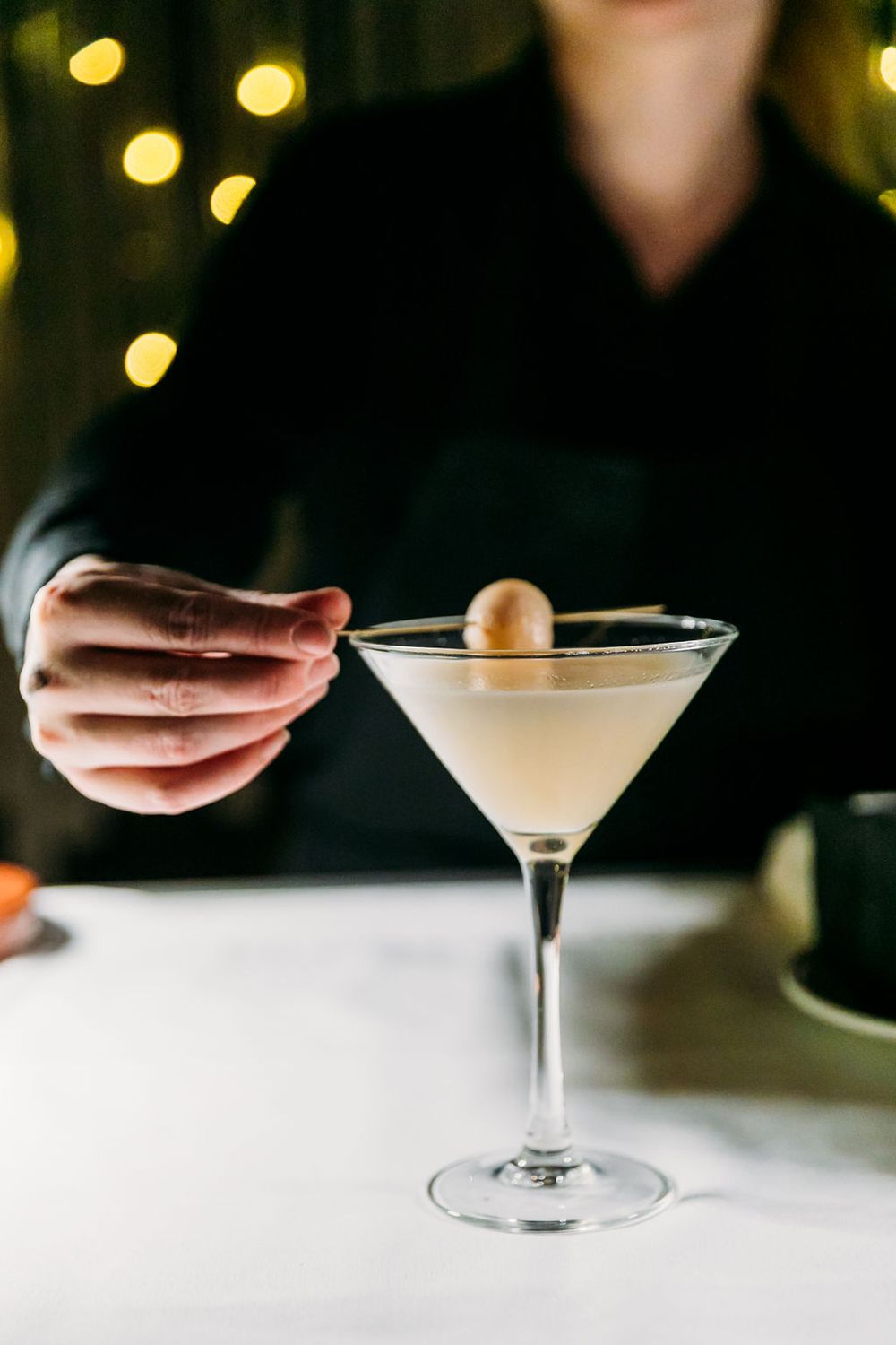 A creamy martini cocktail garnished with a lychee being served on a white tablecloth with bokeh lights in background.