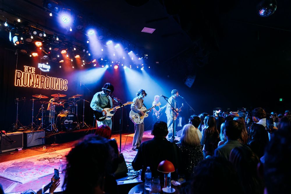 Musicians perform on stage at a dimly lit concert venue with blue lighting and silhouetted crowd in foreground.