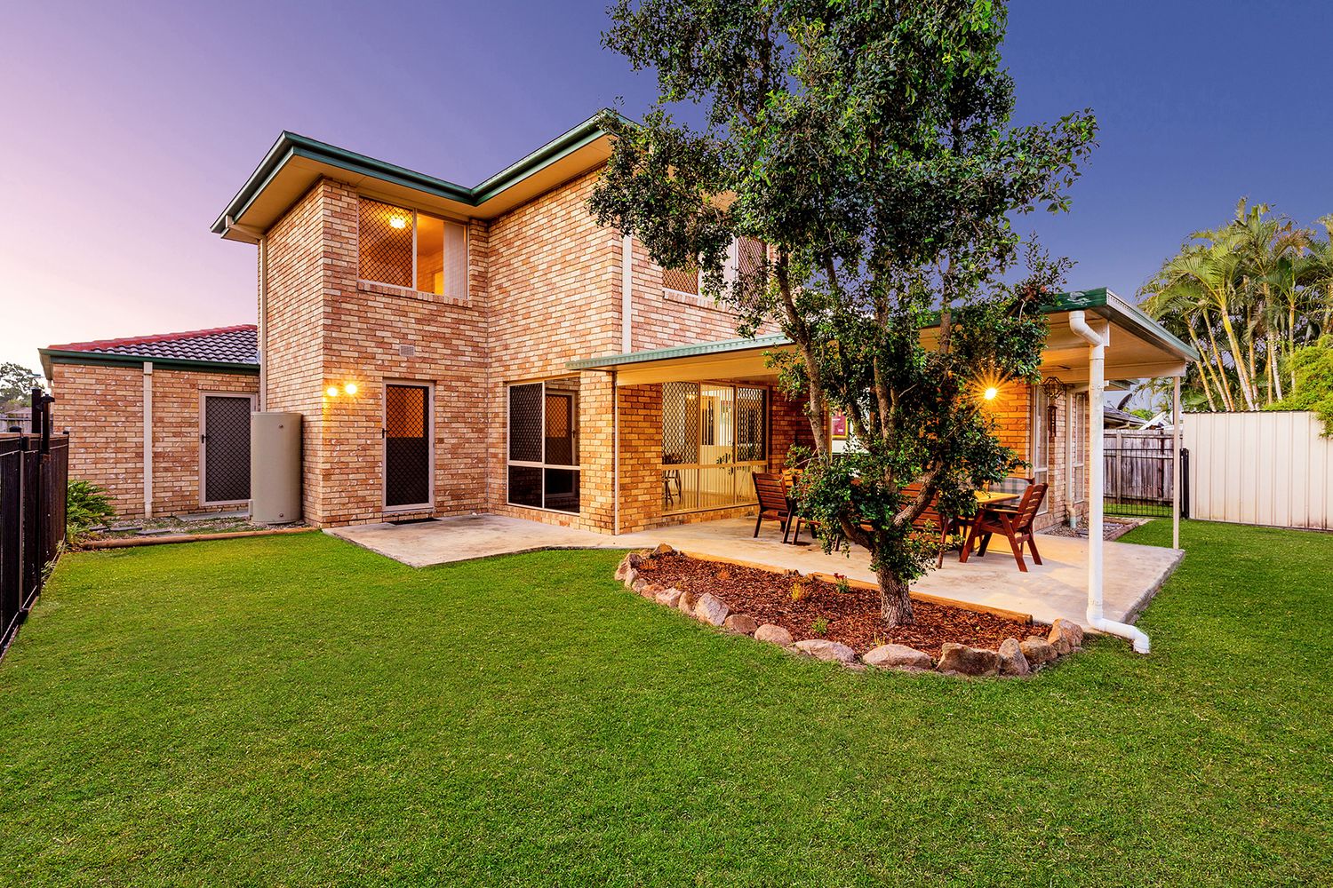 Two-story brick home with covered patio, manicured lawn, and garden tree at dusk.
