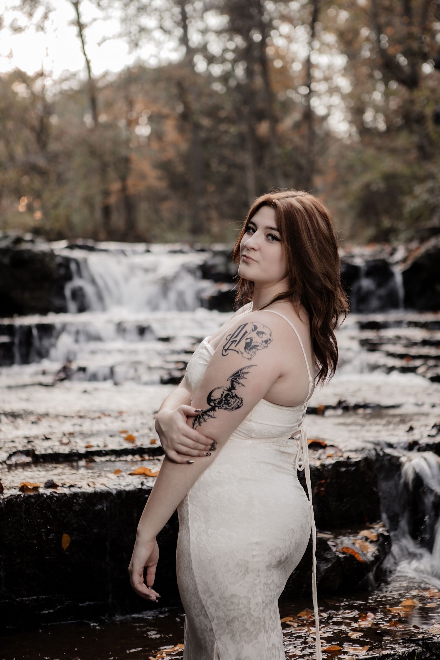 Artistic maternity photoshoot by a waterfall in autumn with fallen leaves and rocks creating a moody atmosphere.