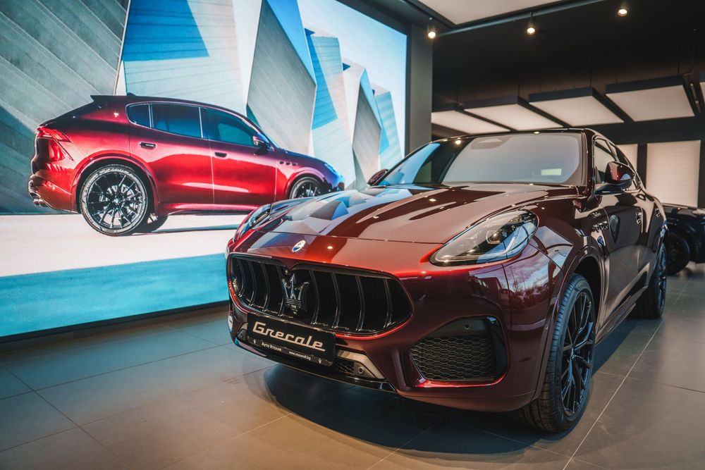 A sleek burgundy Ferrari sports car is displayed in a modern showroom with blue accent lighting and reflective floors.
