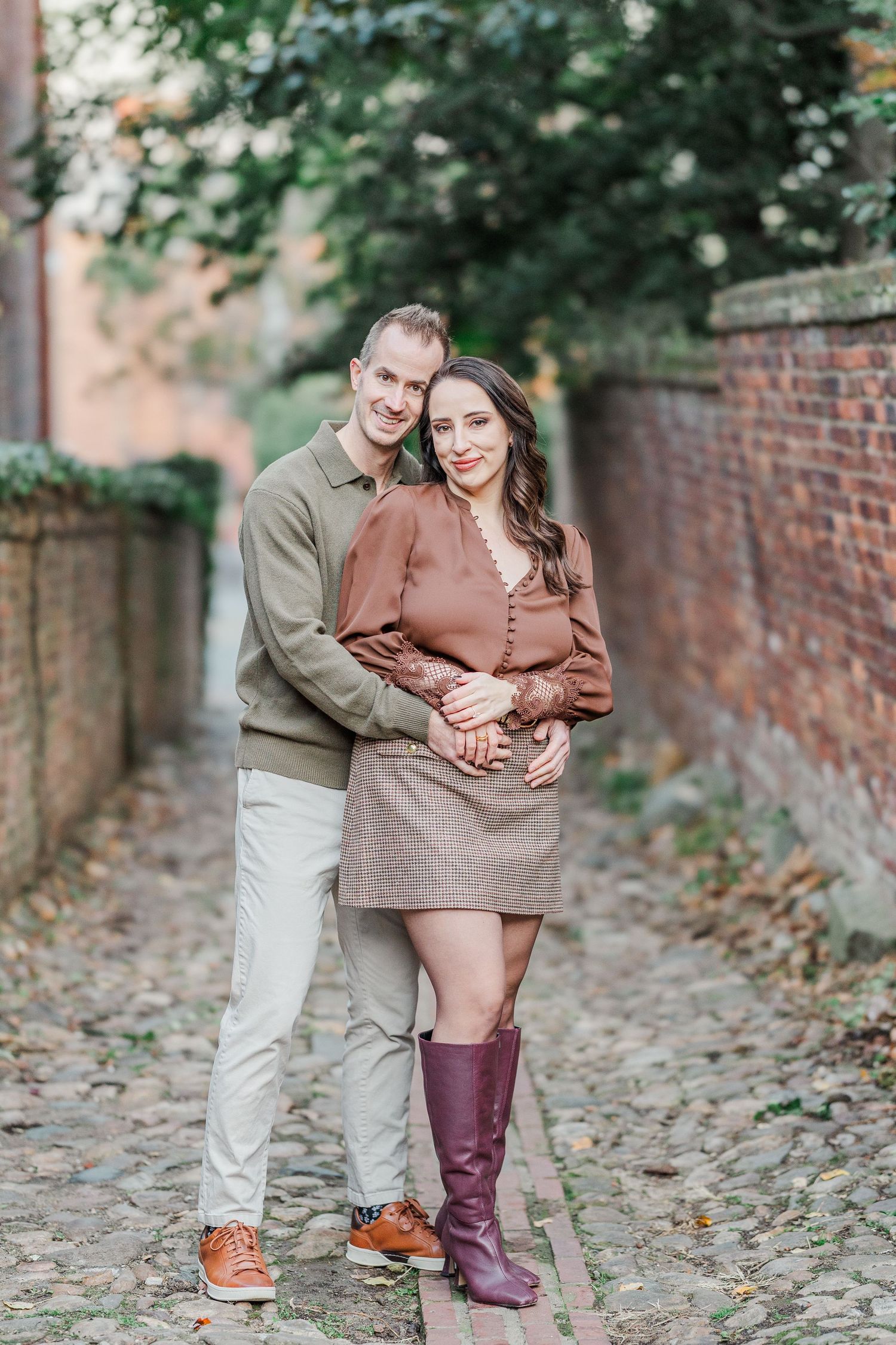 A couple shares tender moments on a cobblestone path near a brick wall surrounded by autumn foliage.