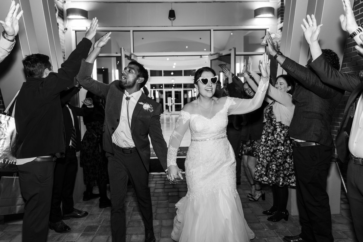 Bride and groom wearing sunglasses celebrate their grand exit through a cheering crowd in black and white.
