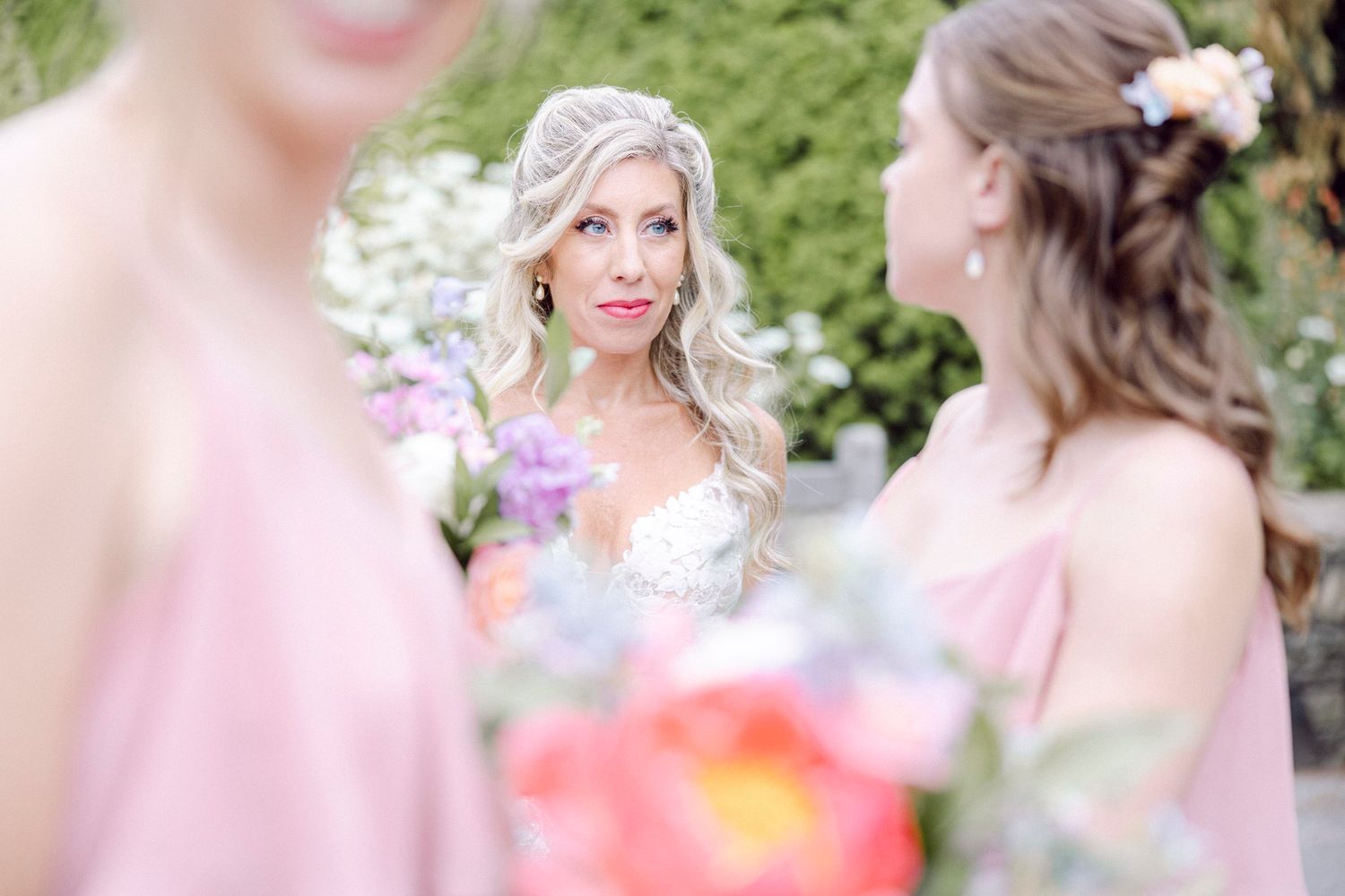 A group of bridesmaids in blush pink dresses gather together in a garden setting for candid wedding photos.