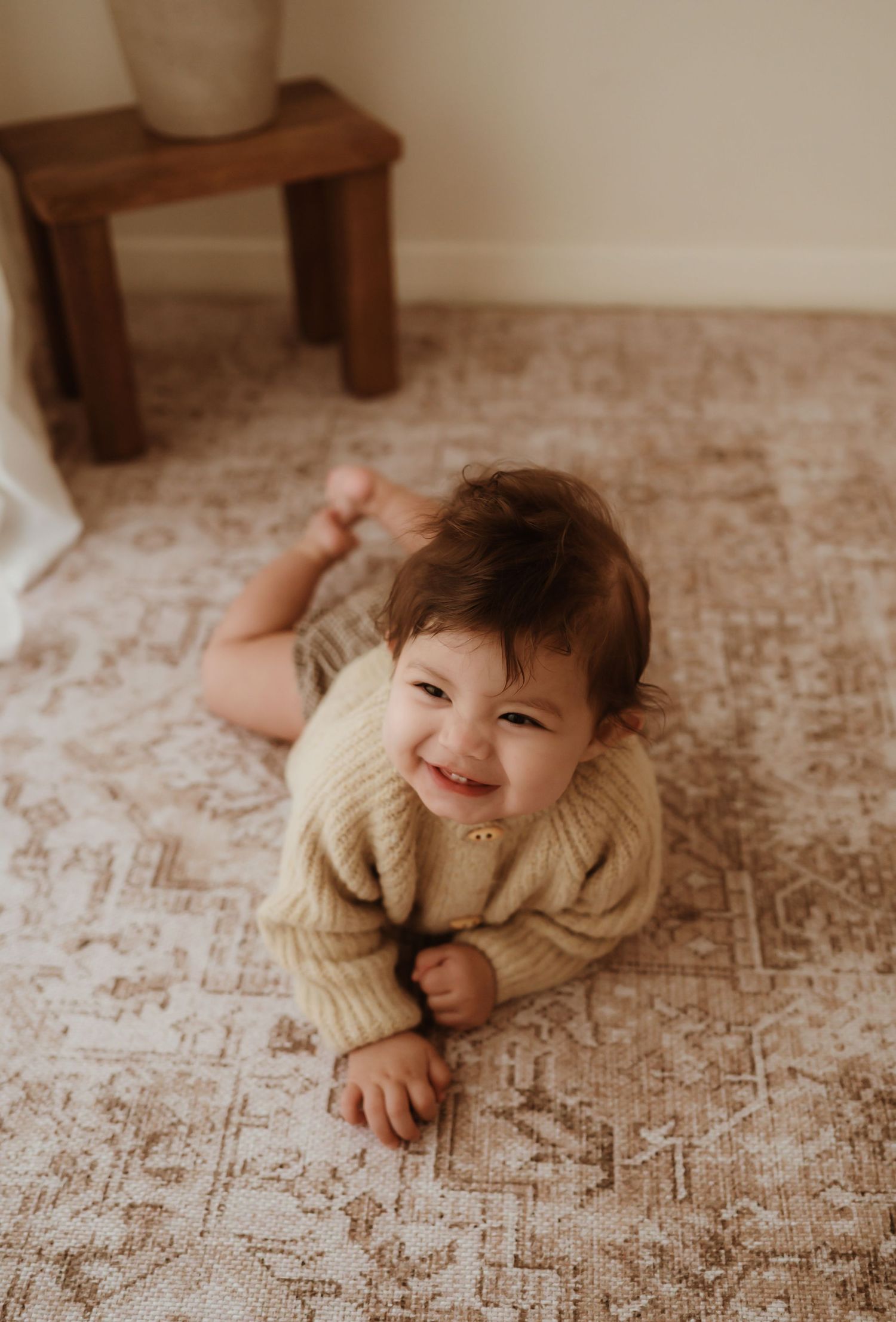 Happy baby in white knit onesie playing with wooden toys on textured rug.