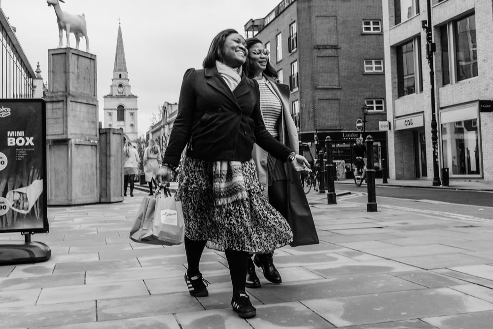 A person in a long coat and patterned skirt walks down a city street with a church spire visible in the background.