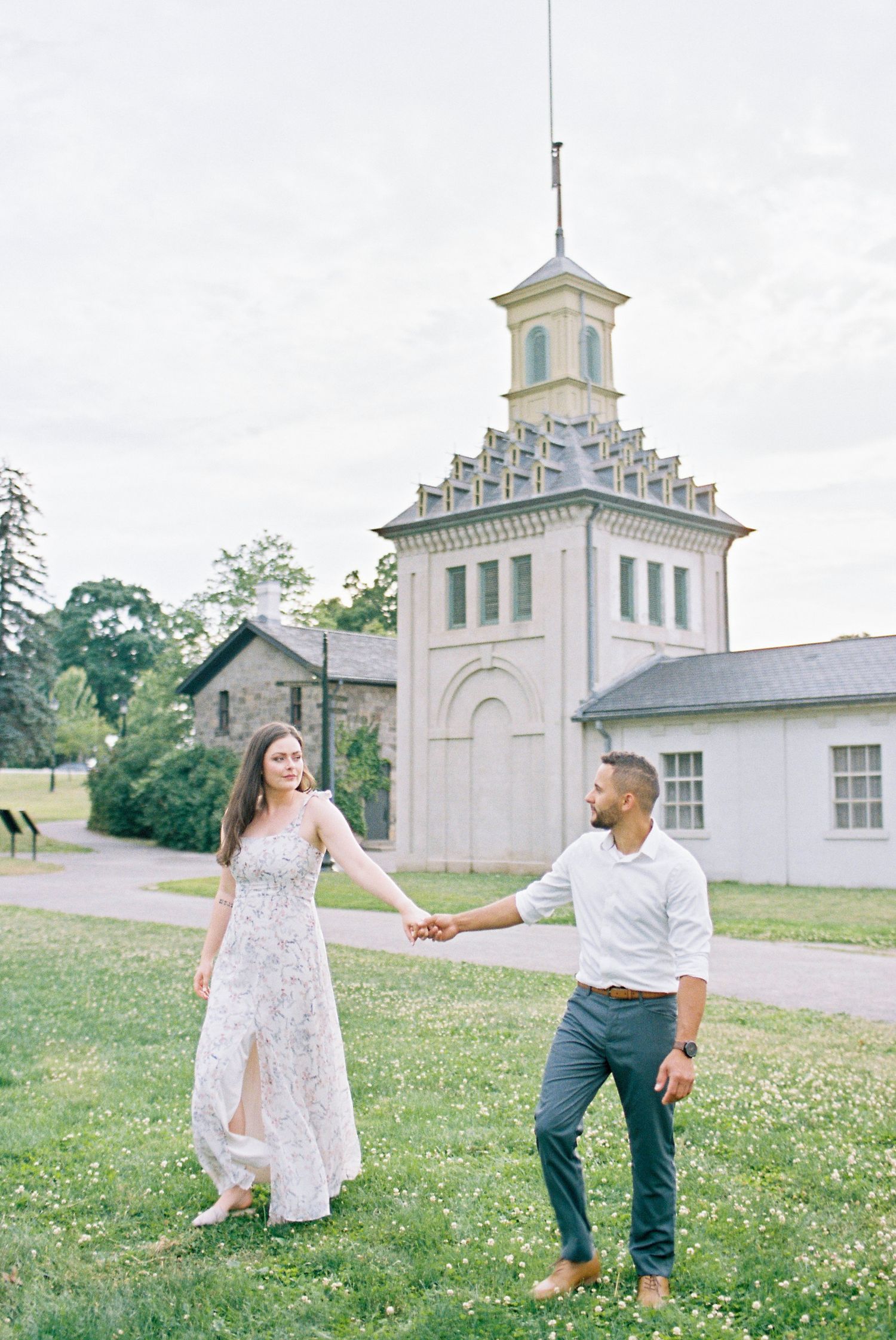 A couple in a meadow poses playfully near a historic church building with white flowers blooming in the grass.