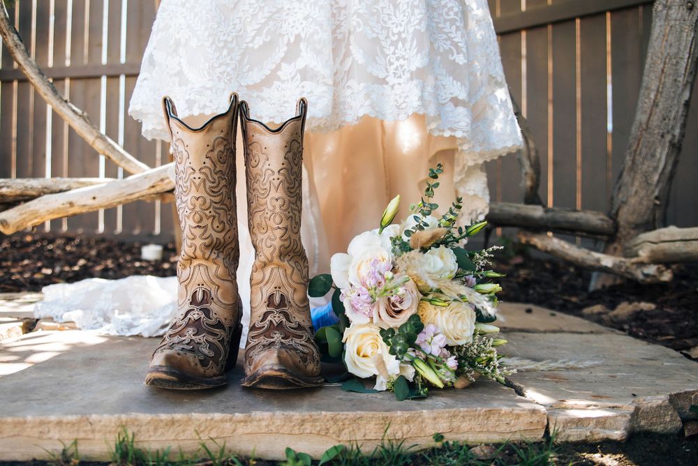A pair of worn brown cowboy boots sits next to a delicate bouquet of pastel flowers and white wedding dress on wooden steps.