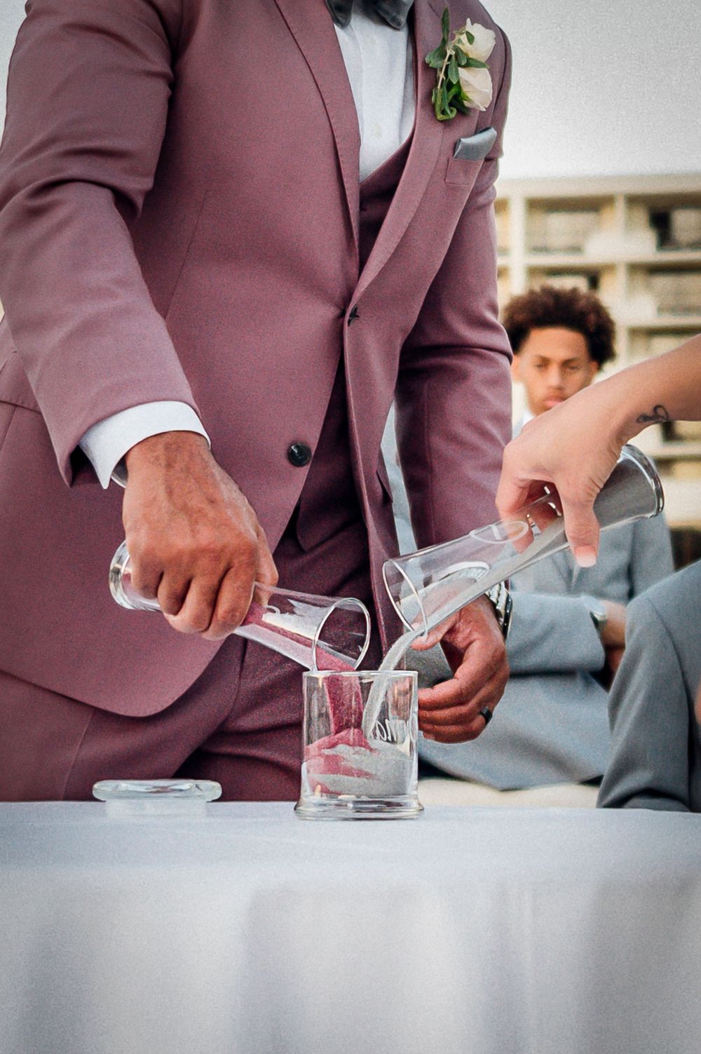 Close-up sequence showing hands in burgundy suit jacket pouring a drink into small glasses at a formal event.