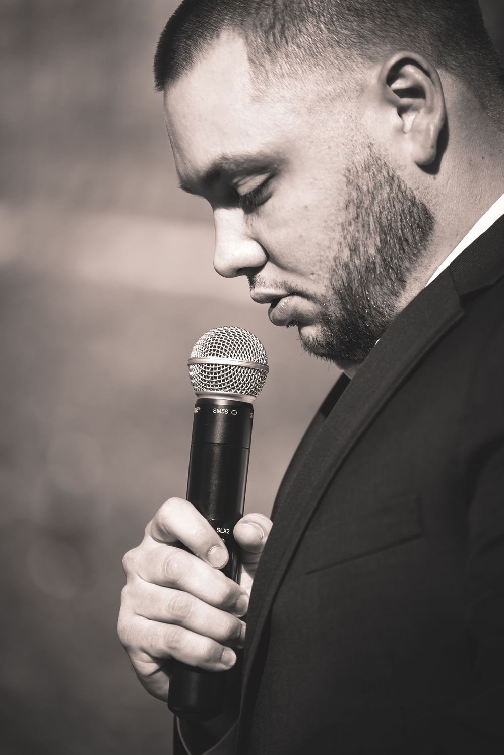 A performer in a black suit holds a microphone in a contemplative pose during a monochrome performance shot.