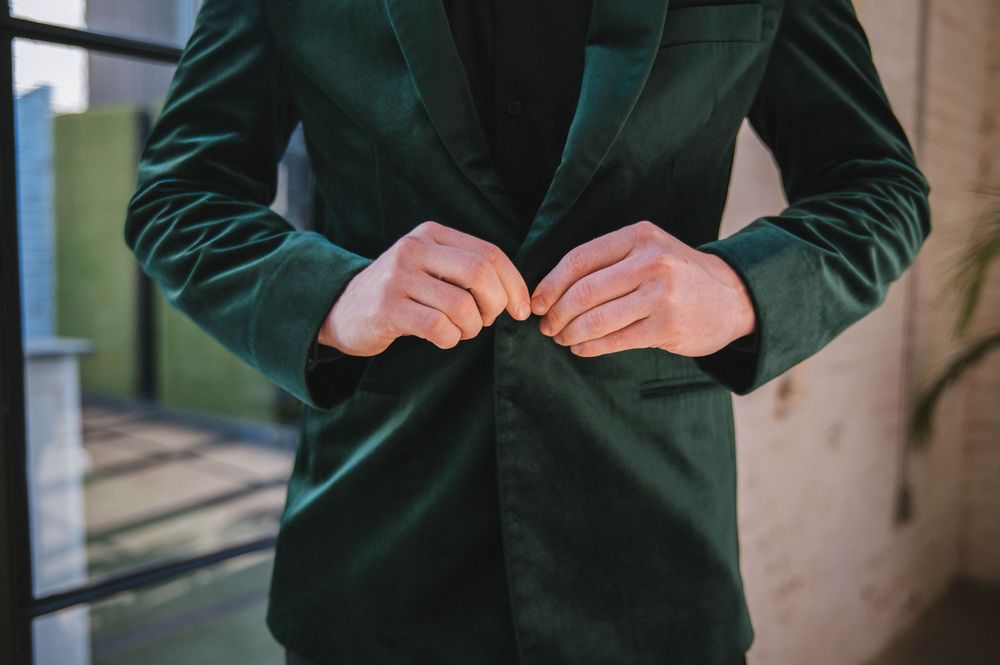 Person in dark green velvet blazer adjusts sleeve cuff detail against neutral background.