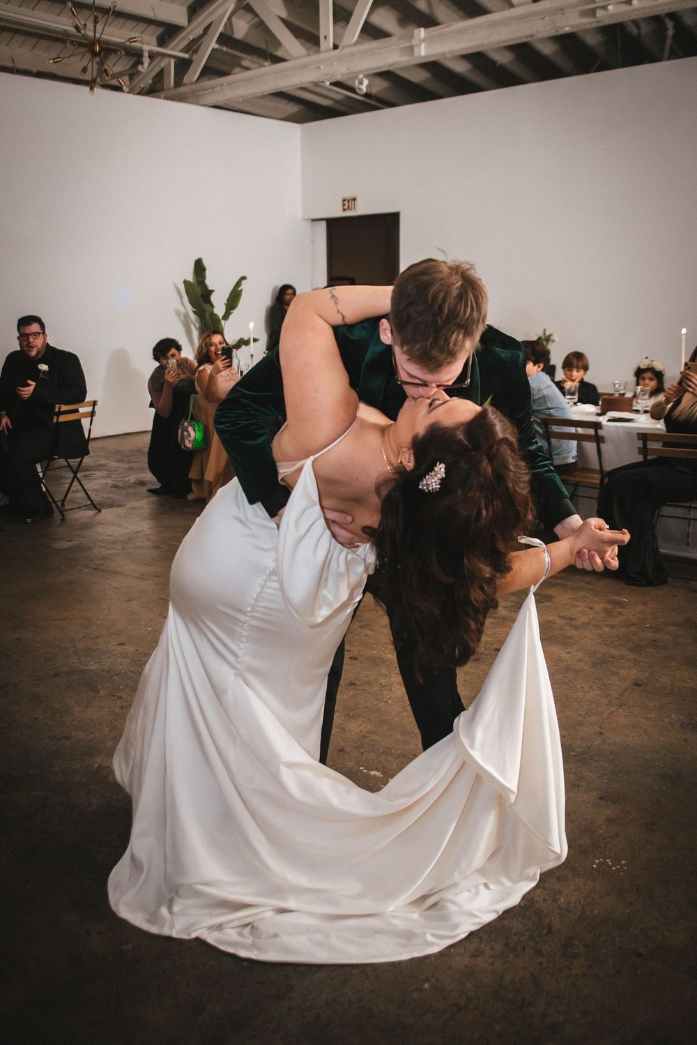 Couples perform dramatic dance dips during a wedding reception in an industrial-style venue with white walls.