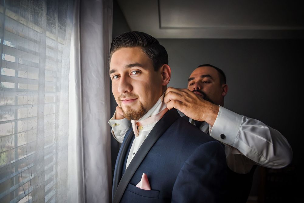 A groom in a dark suit adjusts his tie while standing in a room with window light streaming in.