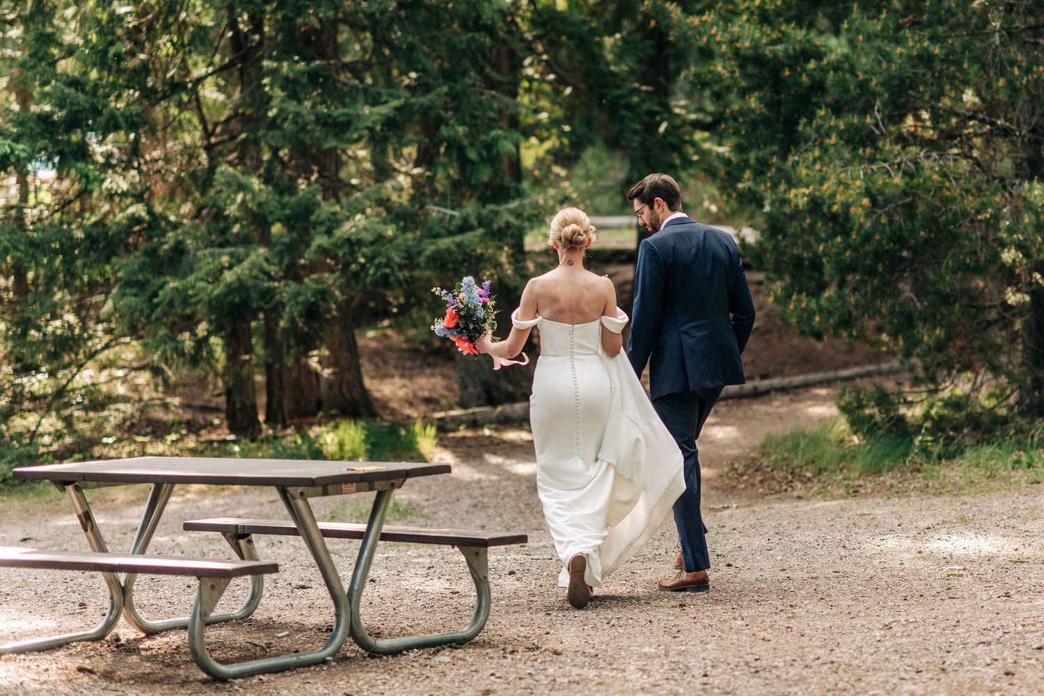 Wedding party relaxes at rustic wooden picnic tables surrounded by trees and greenery.