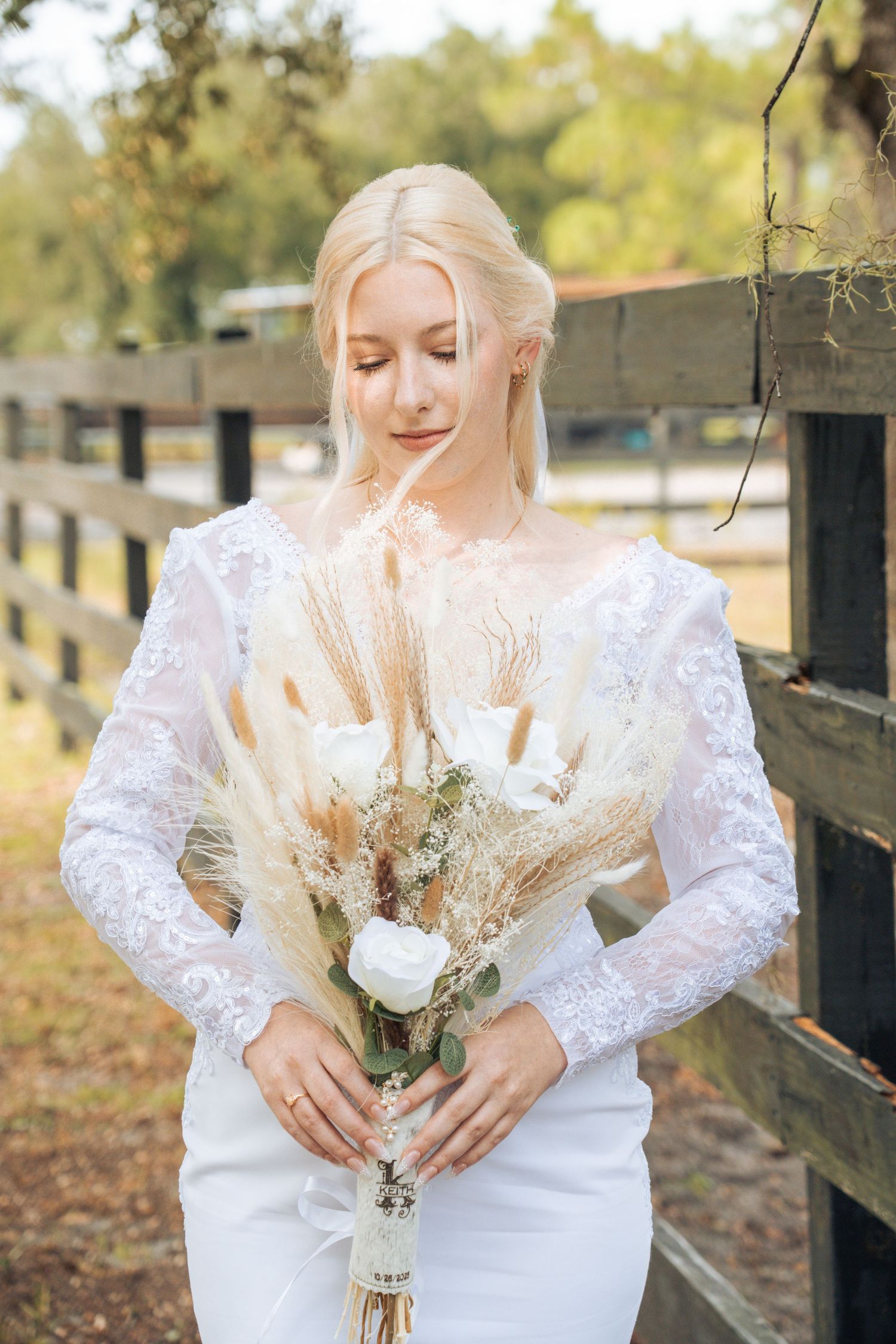 A bride in a white lace dress holds a delicate bouquet of dried flowers and pampas grass against a rustic fence backdrop.