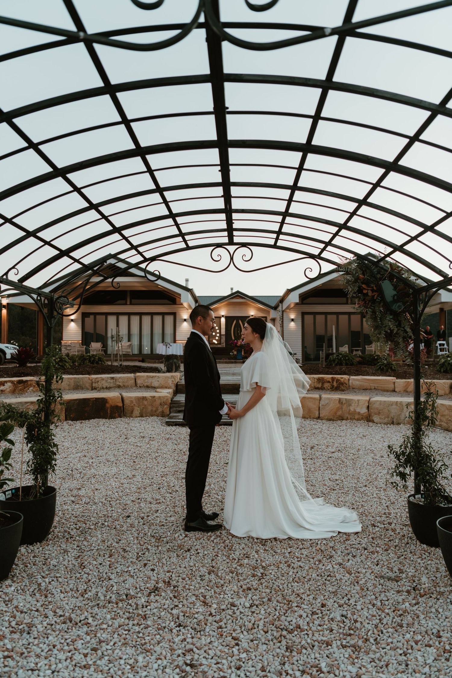 Bride and groom stand facing each other under an elegant arched iron pergola on a gravel path at dusk.