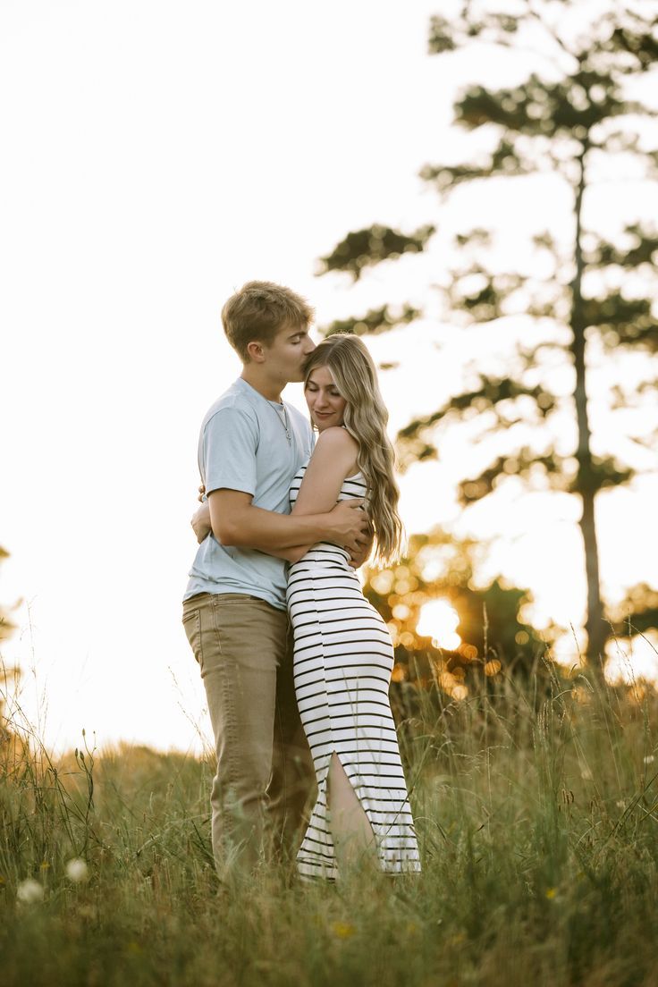 A romantic couple embraces in a grassy field at sunset with tall pine trees silhouetted in the background.