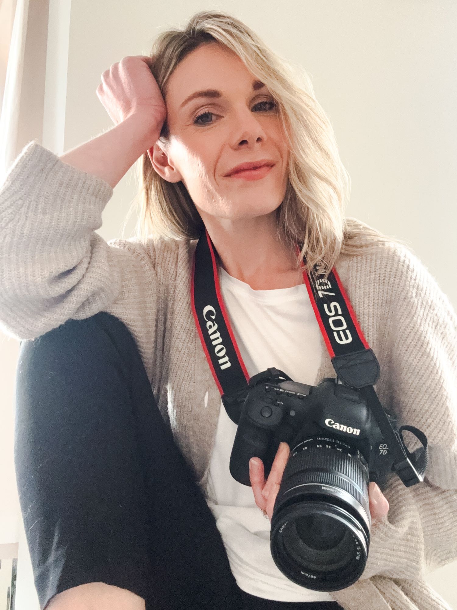 A photographer in a cream sweater poses playfully with a professional DSLR camera in a bright setting.