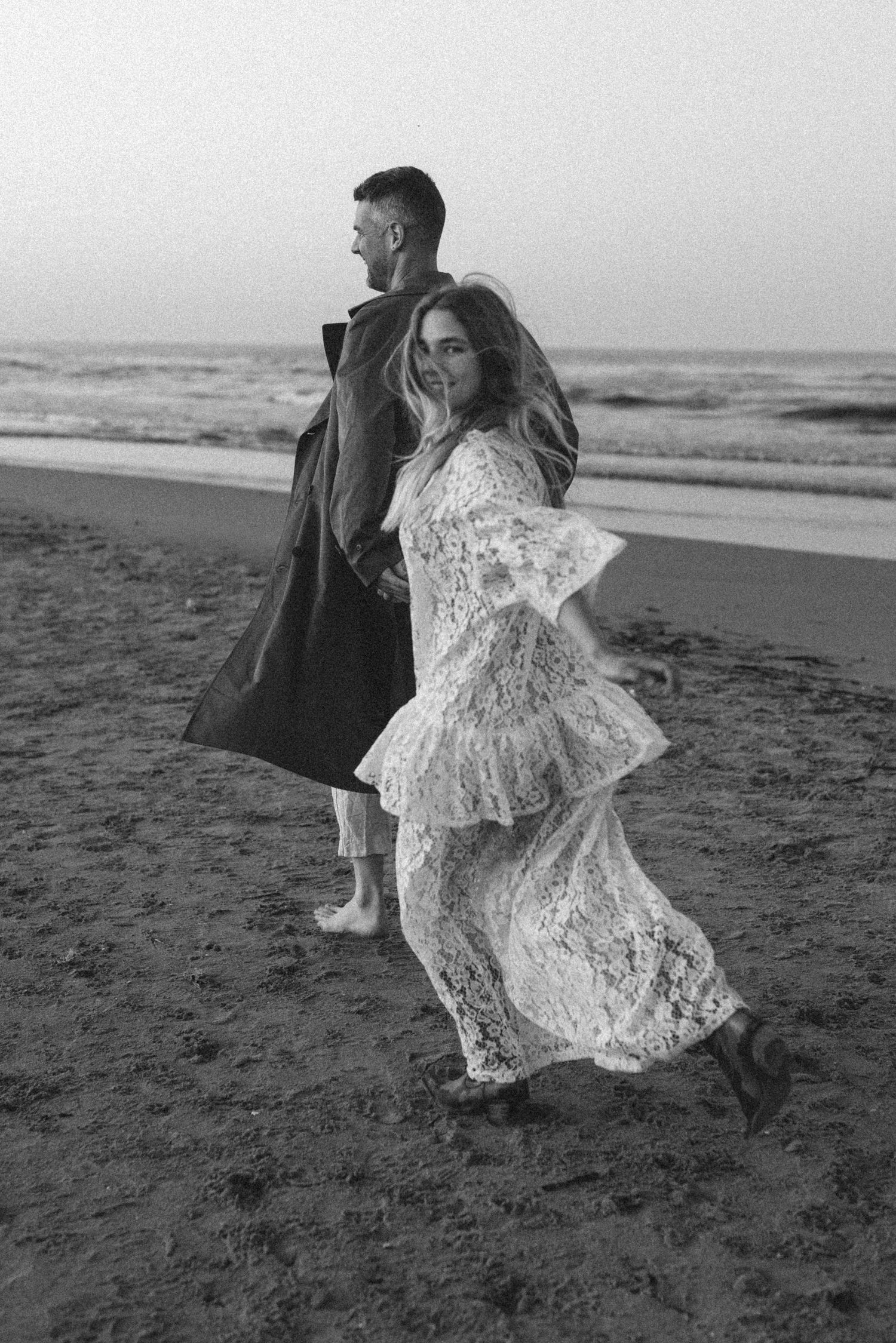 A couple in formal attire runs playfully along a misty beach shore in black and white photography. Javea photographer