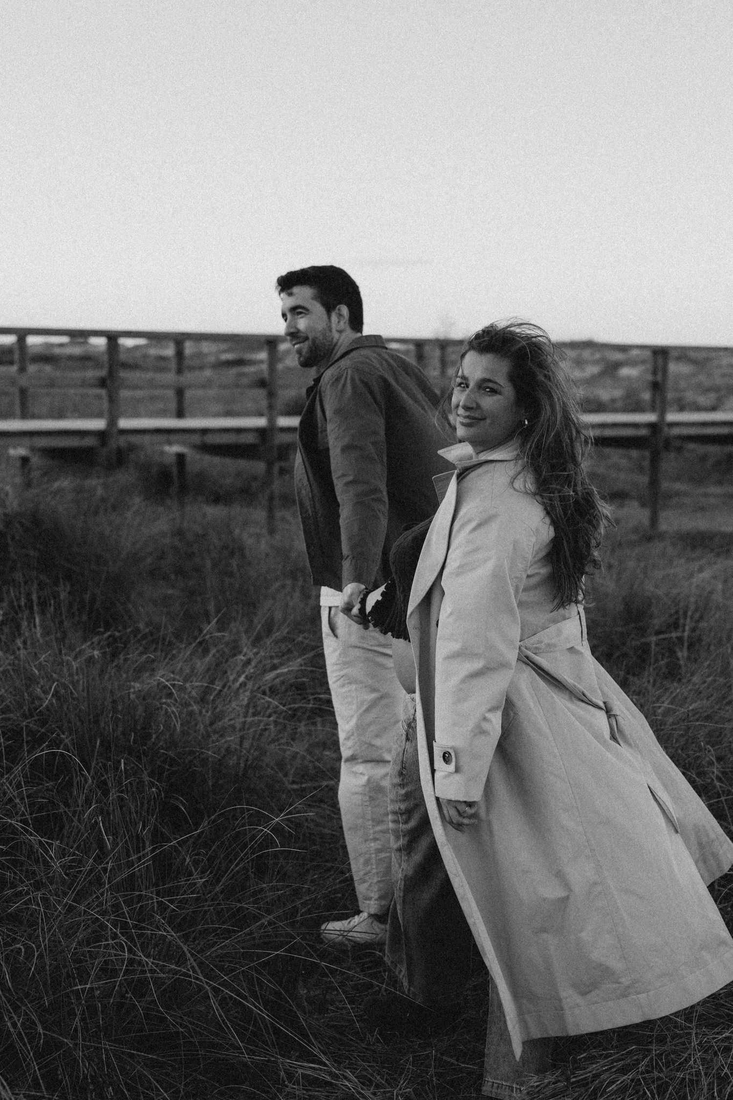 A couple in casual outerwear stands together in a grassy field on an overcast day, captured in black and white. Javea photographer