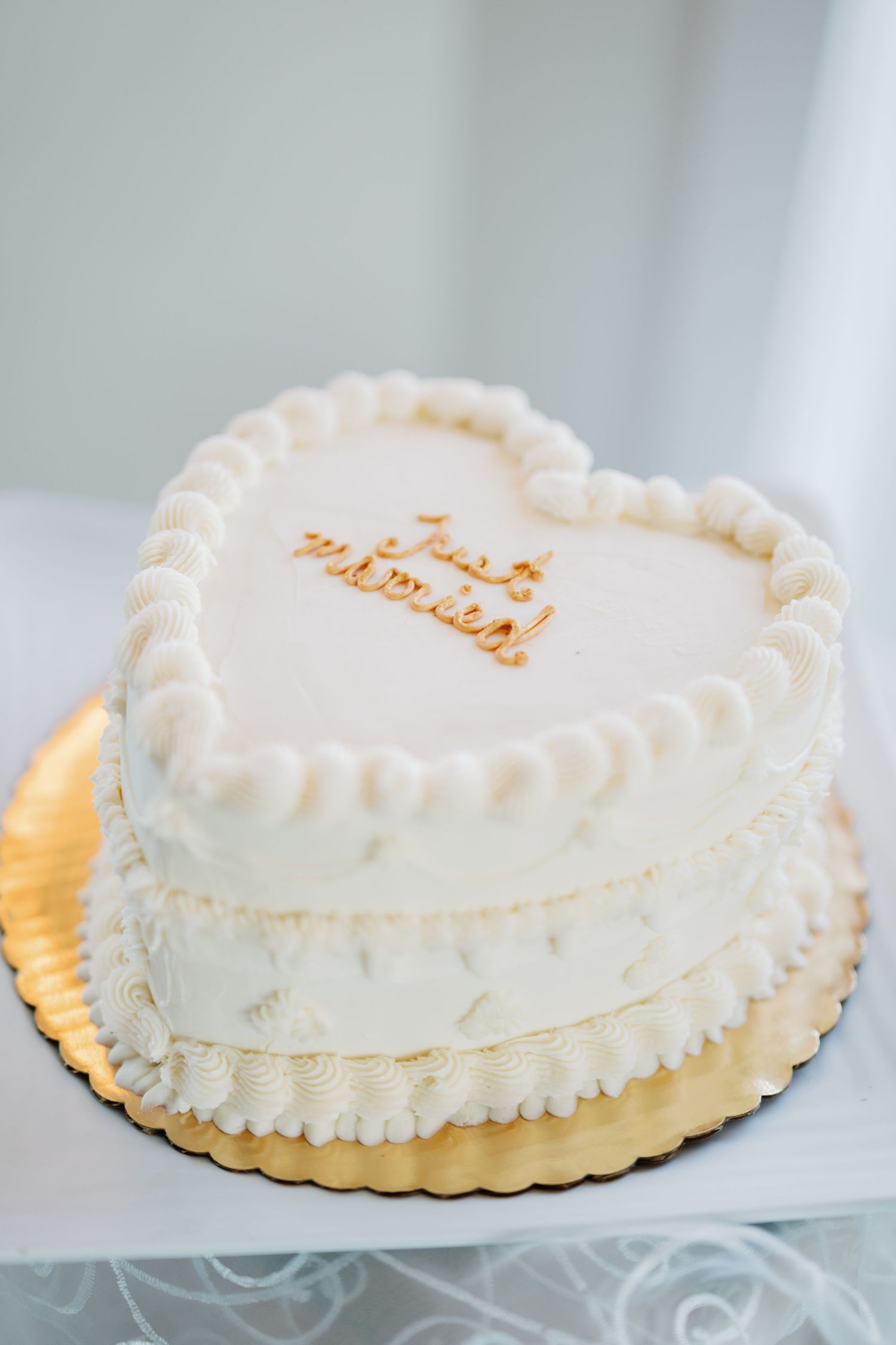 A heart-shaped white wedding cake with delicate piped frosting details and gold text sits on a gold cake board.