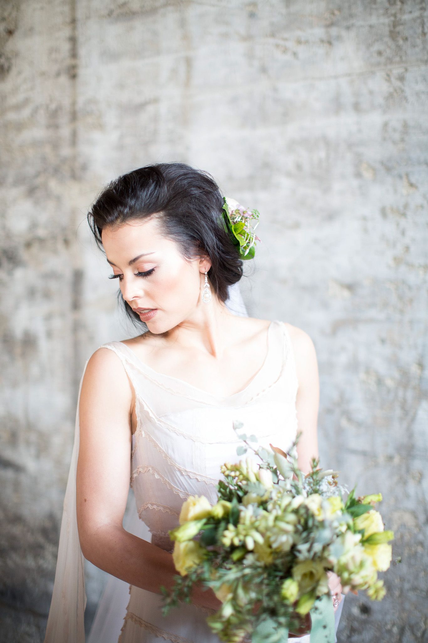 Elegant dark-haired bride in a lace gown holding a green and yellow bouquet against a concrete wall.