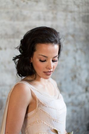 Beautiful dark-haired bride in a delicate lace wedding dress looking down gracefully against a grey wall.