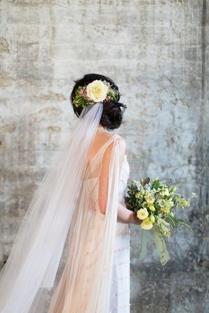 Back view of bride with floral hair crown and veil in a lace gown holding a yellow bouquet.