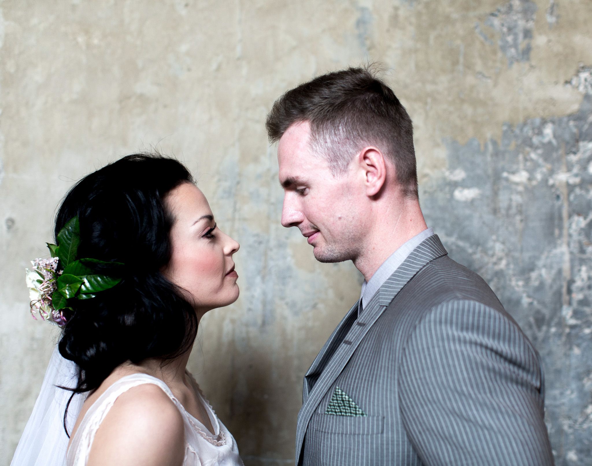 Bride and groom facing each other intimately against rustic concrete wall.