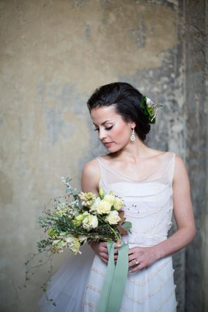 Elegant bride in white dress holding green and white bouquet against wall.