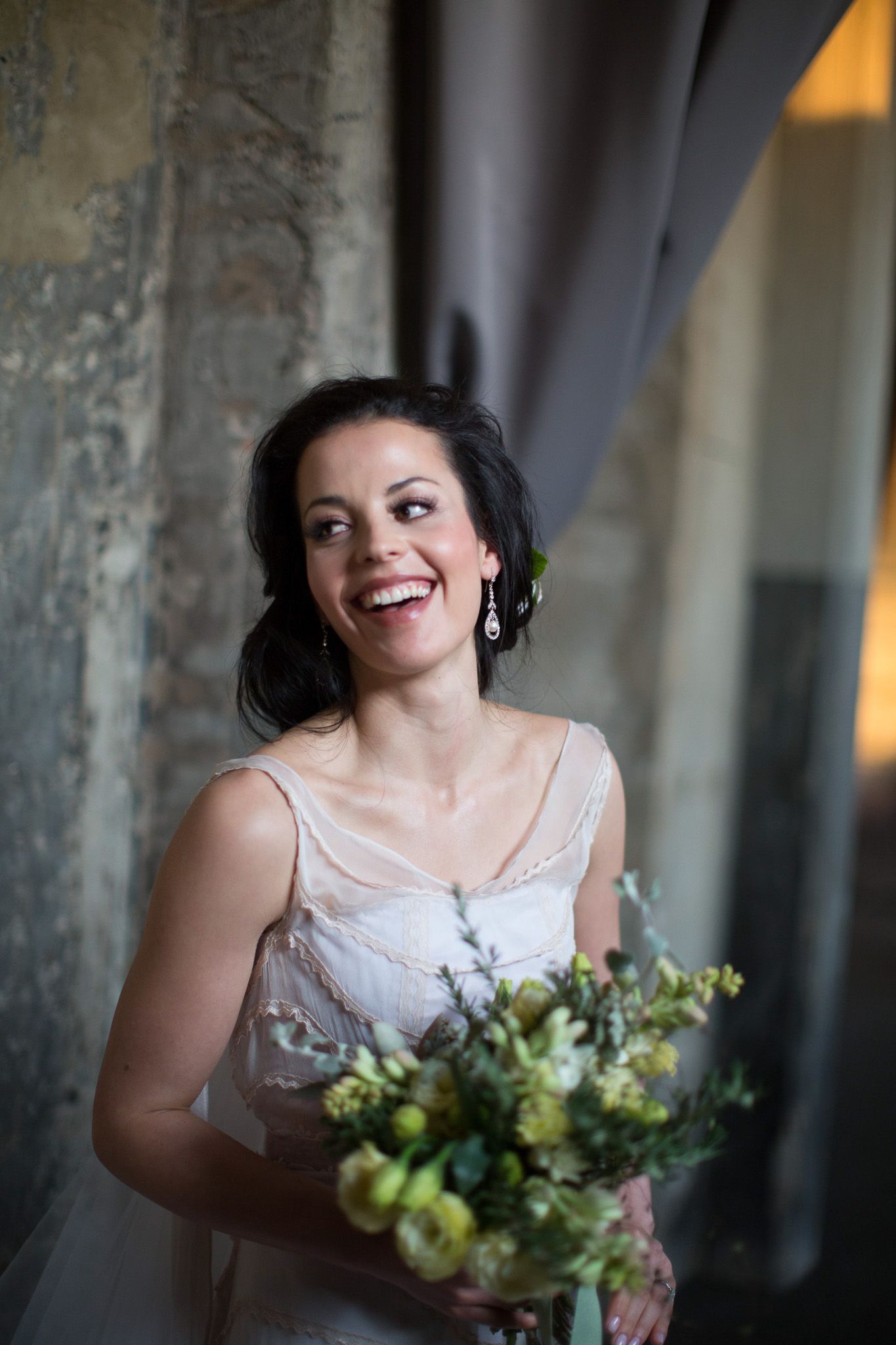 Smiling bride in white dress holding green and yellow bouquet laughing joyfully indoors.