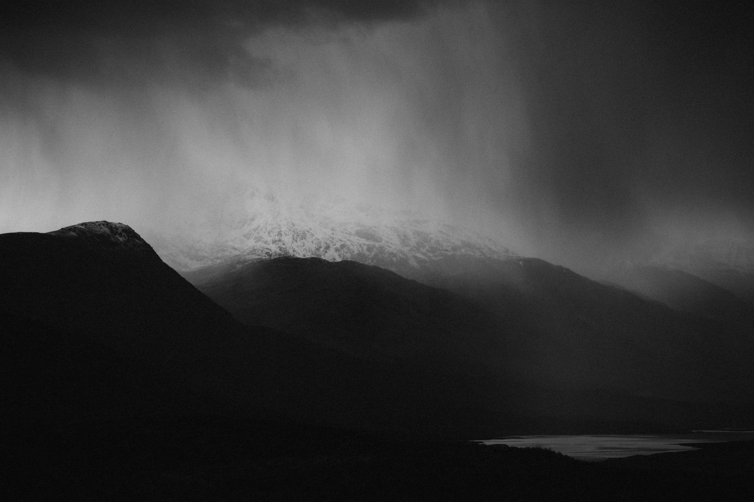 Moody black and white landscape of mountains with rain and low clouds over a distant lake.