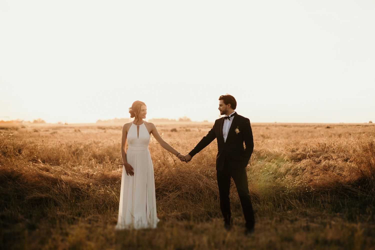 A bride and groom hold hands while standing in a golden field during sunset.